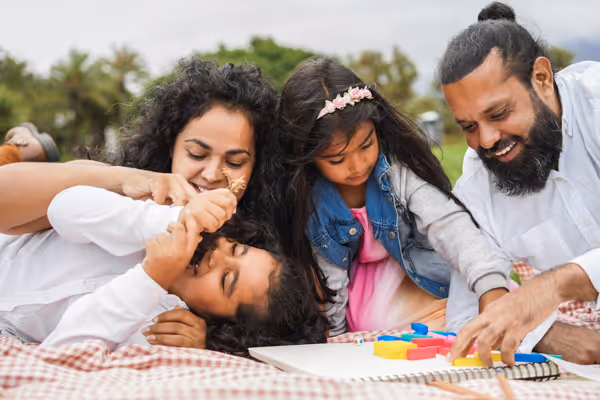 A happy family of four playing together on a blanket outdoors with a colorful board game.