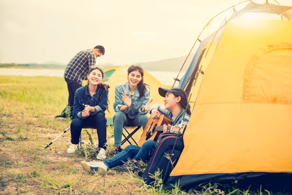 Group of friends camping outdoors with a yellow tent, one playing guitar while others sit and smile.