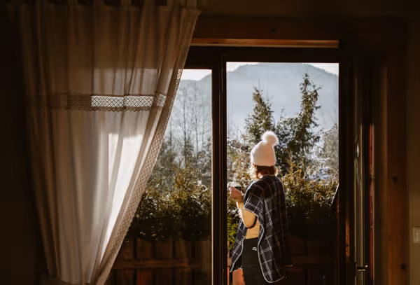 Person wearing a white woolen hat and plaid shirt standing on a balcony looking at trees and mountains.