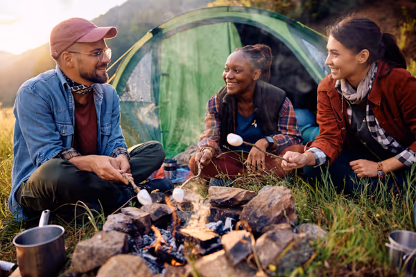 Three friends sitting around a campfire roasting marshmallows near a green tent at sunset.