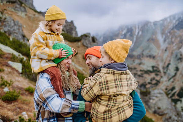 Smiling family of four wearing winter hats and jackets, enjoying time together outdoors in a mountainous landscape.