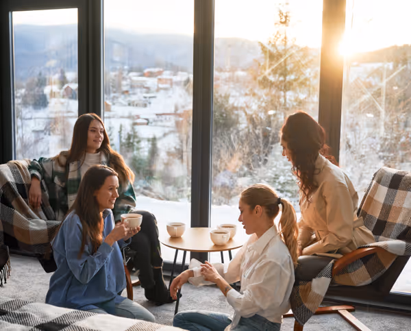 Four women sitting and enjoying coffee together in a cozy room with large windows and a scenic snowy mountain view.