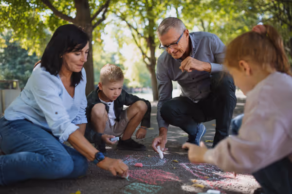 Family of four drawing with colorful chalk on sidewalk under leafy trees in a park.