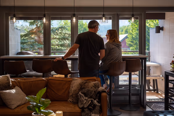 A man and woman sitting on bar stools facing each other and smiling in a cozy room with large windows overlooking green trees.