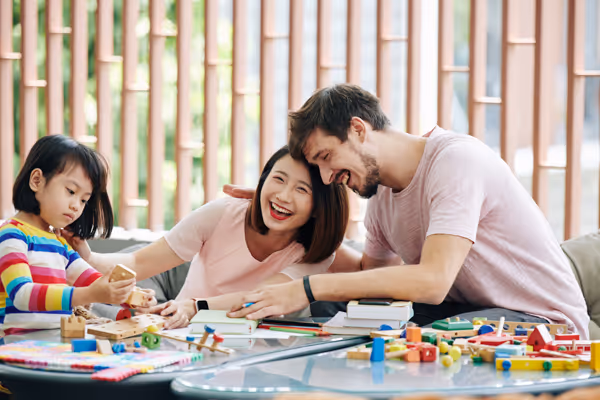 Smiling family with a young girl playing with wooden toys on a glass table indoors.