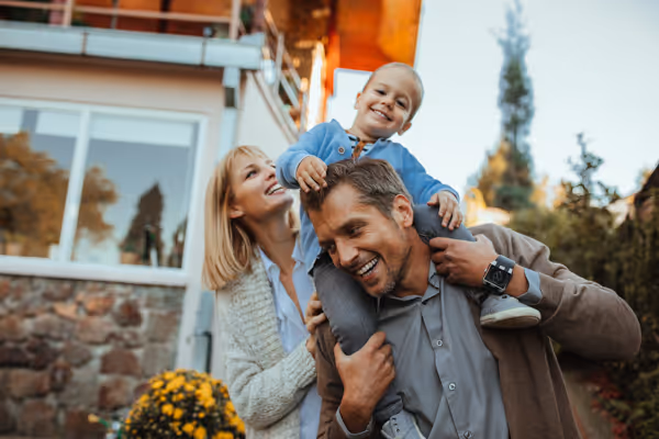 Smiling father carrying a happy young boy on his shoulders with a woman standing beside them outdoors near a house.