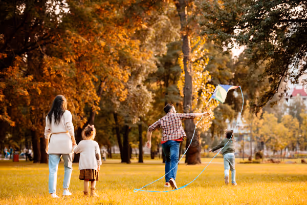 A woman and three children flying a kite together in a sunlit autumn park with trees in the background.